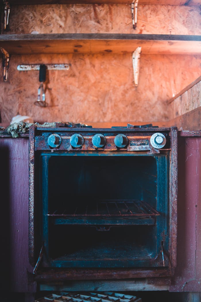 gallery-4 Old broken metal gas stove with rusty parts placed in old kitchen under wooden shelves